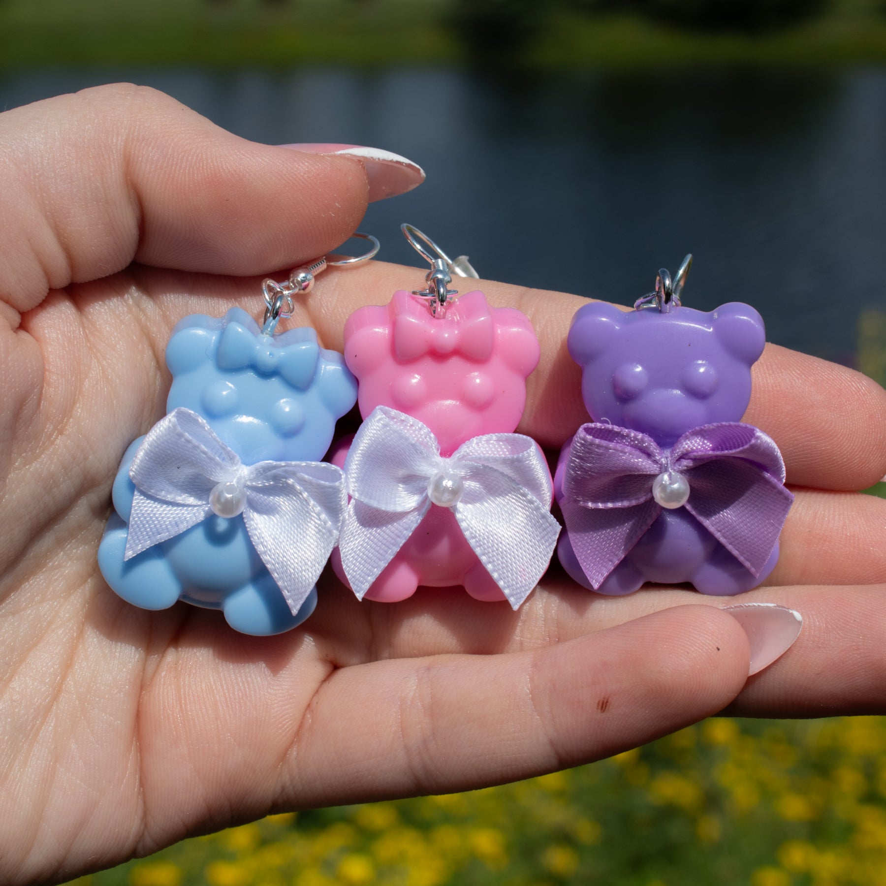 Three colorful bear-shaped earrings with bows held in a hand against a natural background.