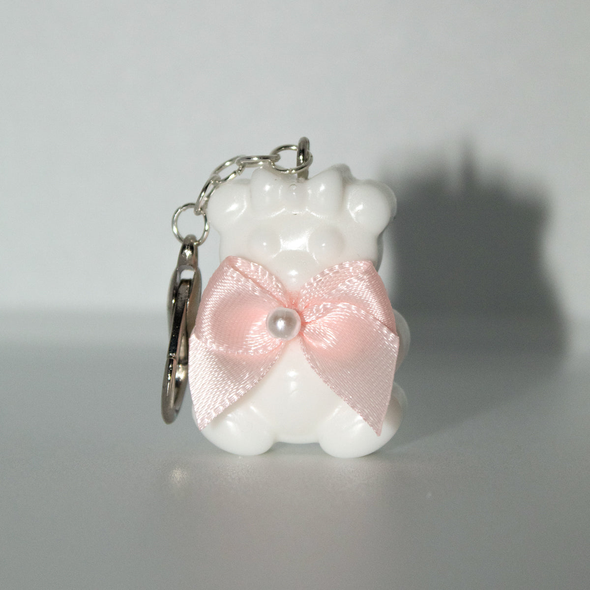 White bear-shaped keychain with a pink bow and pearl on a gray background